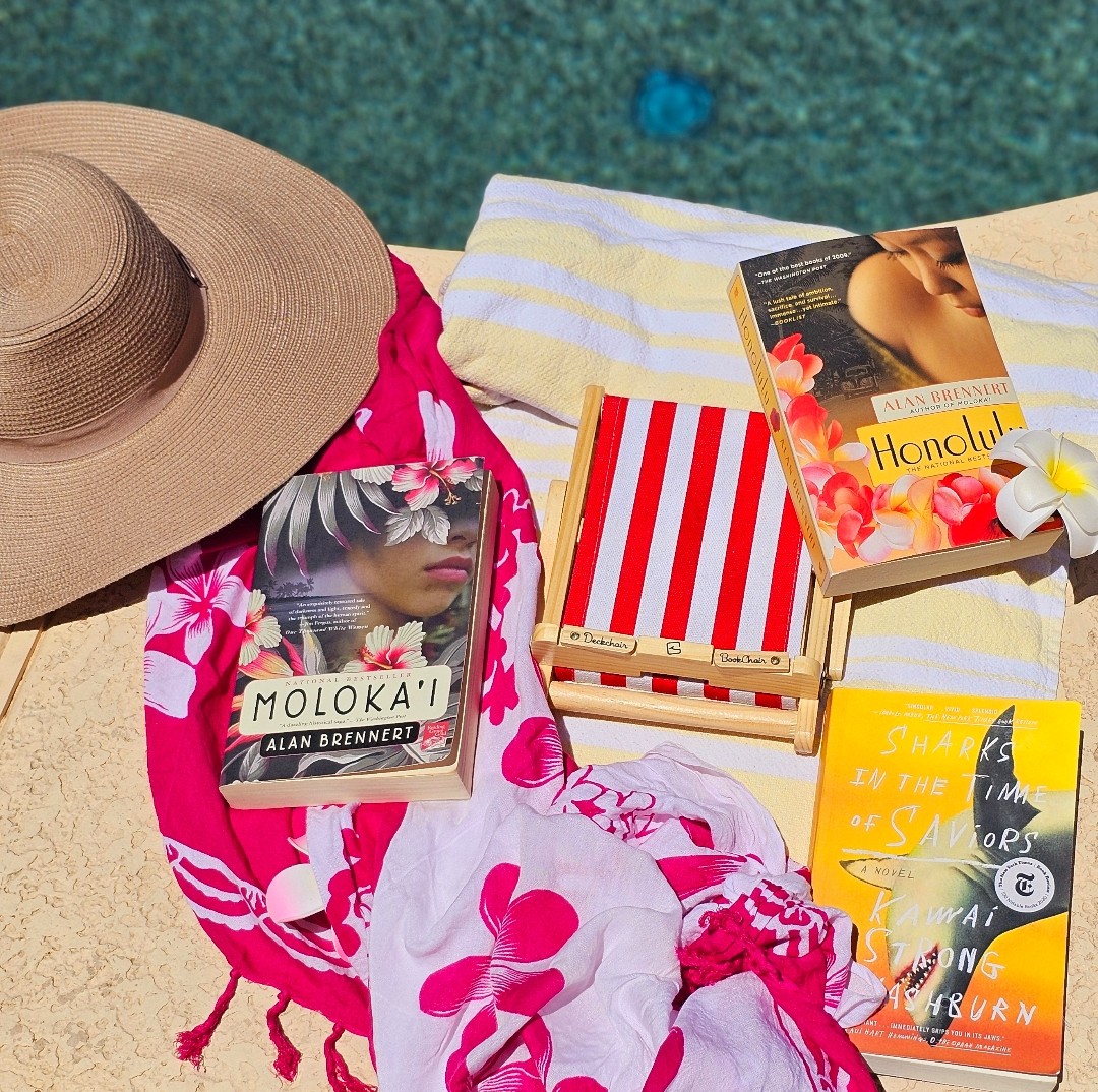 Several Hawaiian themed books are lying on top of a beach towel and a striped beach chair the size of a paperback book. Book titles include: Moloka'i, Honolulu, and Sharks in the Time of Saviors.