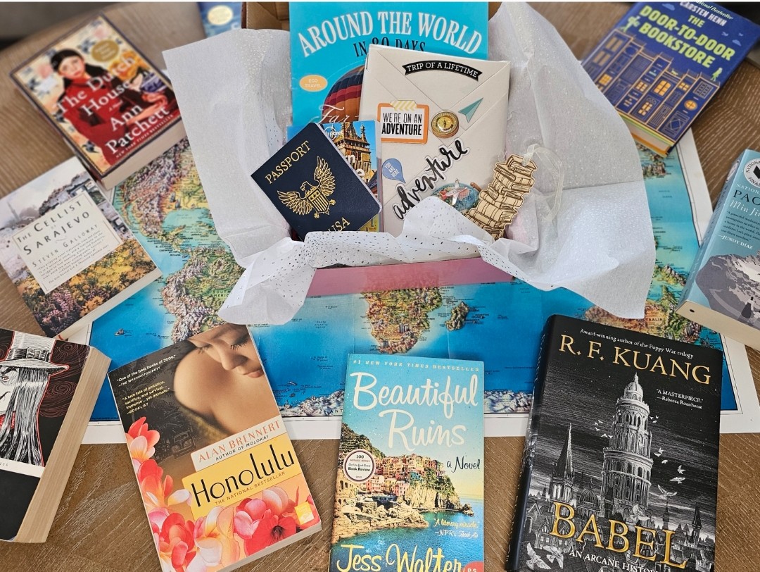 A pink box sits on top of a world map. Inside the box is a wrapped book stamped like a passport, a reading passport journal, a guidebook, a wooden bookmark shaped like stacked books, and a passport sticker. Surrouding the box of items are a variety of books from around the world, including: England, Italy, Romania, Bosnia, South Korea, and the Netherlands.