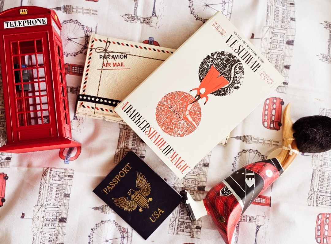 There is a black and red book called A Darker Shade of Magic in the center of the photo next to a red telephone booth, passport book, and a wooden duck dressed like a British guard (Beefeater). 