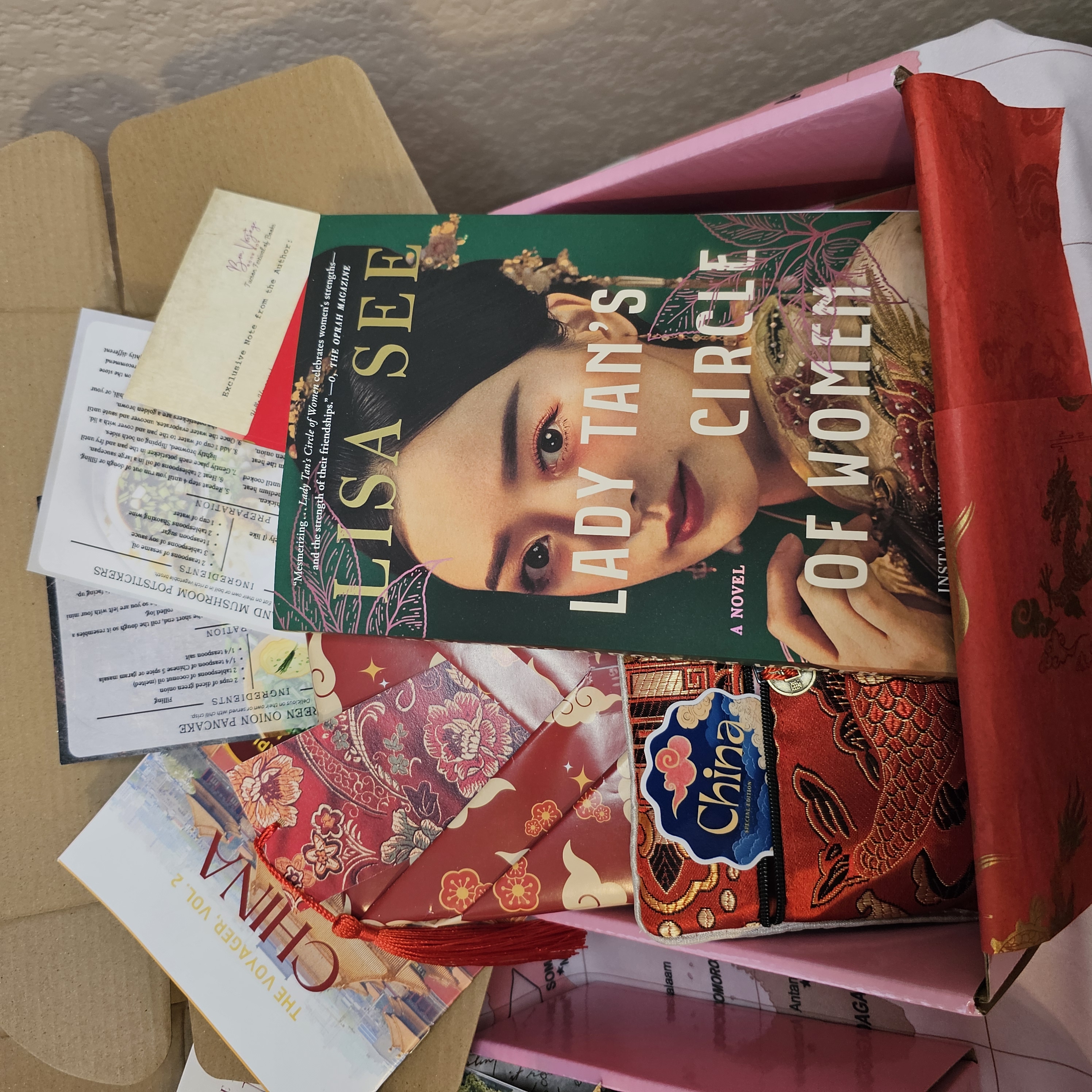 What's inside the Bon Voyage Book box? A pink box sits on top of a world map. Inside the box is a wrapped book stamped like a passport, a reading passport journal, a guidebook, a wooden bookmark shaped like stacked books, and a passport sticker. Surrouding the box of items are a variety of books from around the world, including: England, Italy, Romania, Bosnia, South Korea, and the Netherlands.