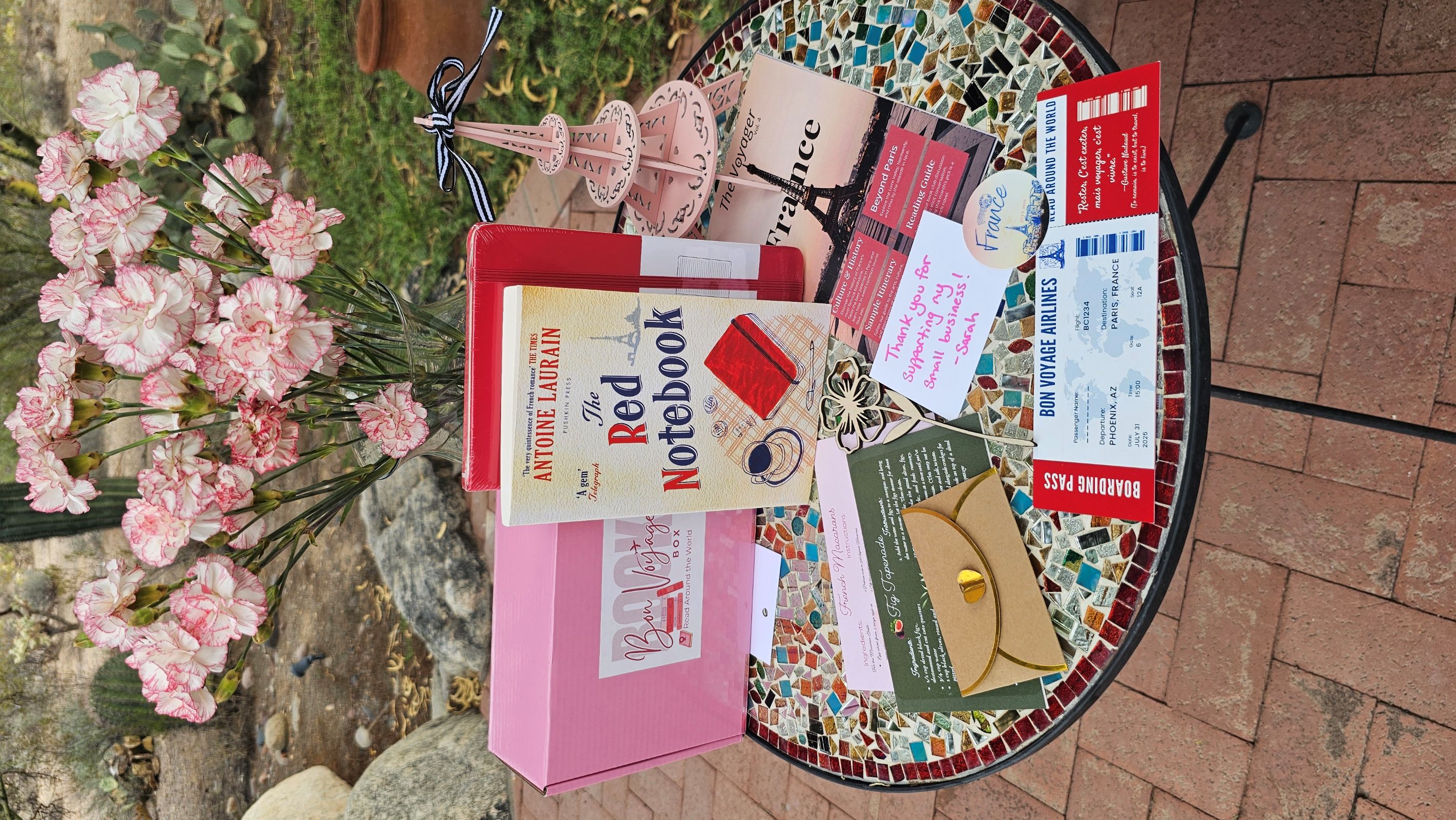 What's inside the Bon Voyage Book box? A pink box sits on top of a world map. Inside the box is a wrapped book stamped like a passport, a reading passport journal, a guidebook, a wooden bookmark shaped like stacked books, and a passport sticker. Surrouding the box of items are a variety of books from around the world, including: England, Italy, Romania, Bosnia, South Korea, and the Netherlands.