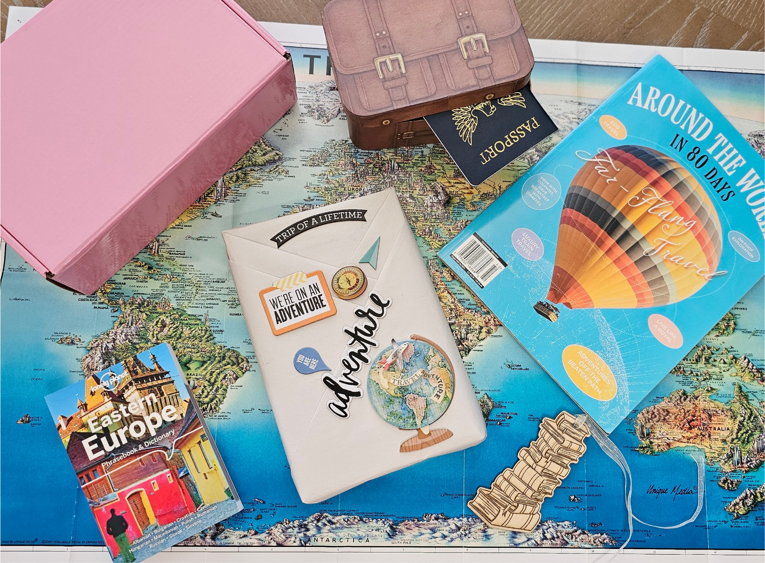 A wrapped book is covered with travel-themed stickers and passport stamps. Next to it is a guidebook to Eastern Europe, a travel magazine, and a passport sticking out of a box that looks like a suitcase. 