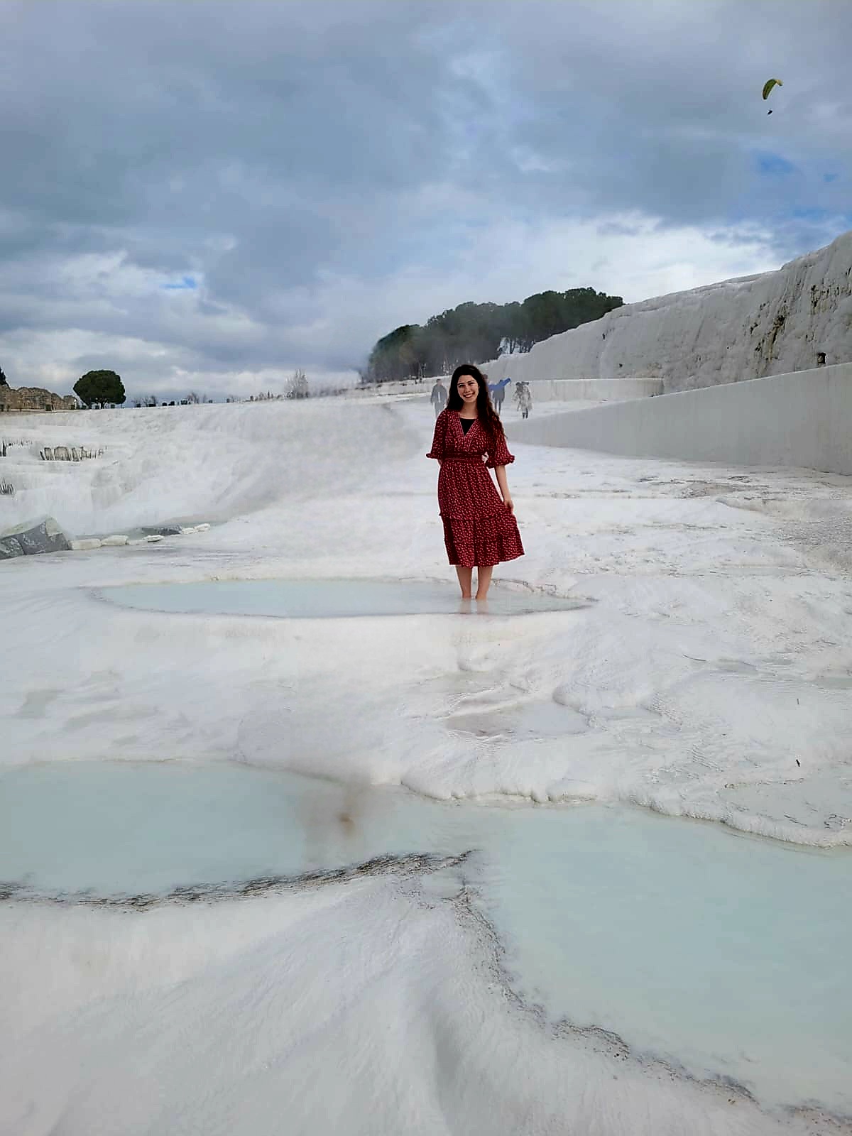 A young woman in a red dress stands in the middle of a salt fields with hot water pools.