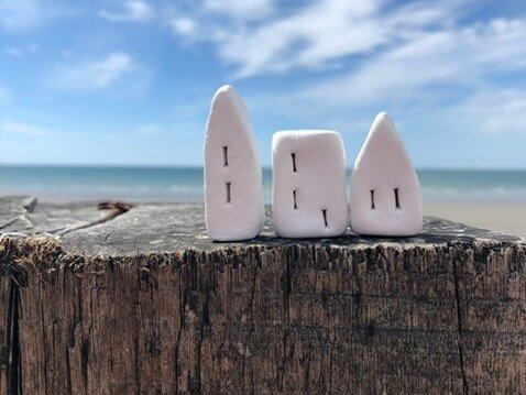 Three tiny clay houses that are painted white, sit on top of a wooden stump. The beach and sea are visible in the background, with bright blue skies.