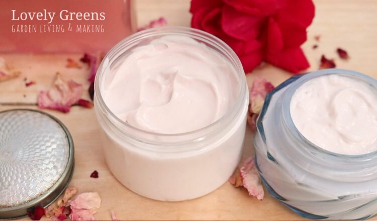 Two jars of a pale pink rose body cream sit atop a wooden table, surrounded by rose petals