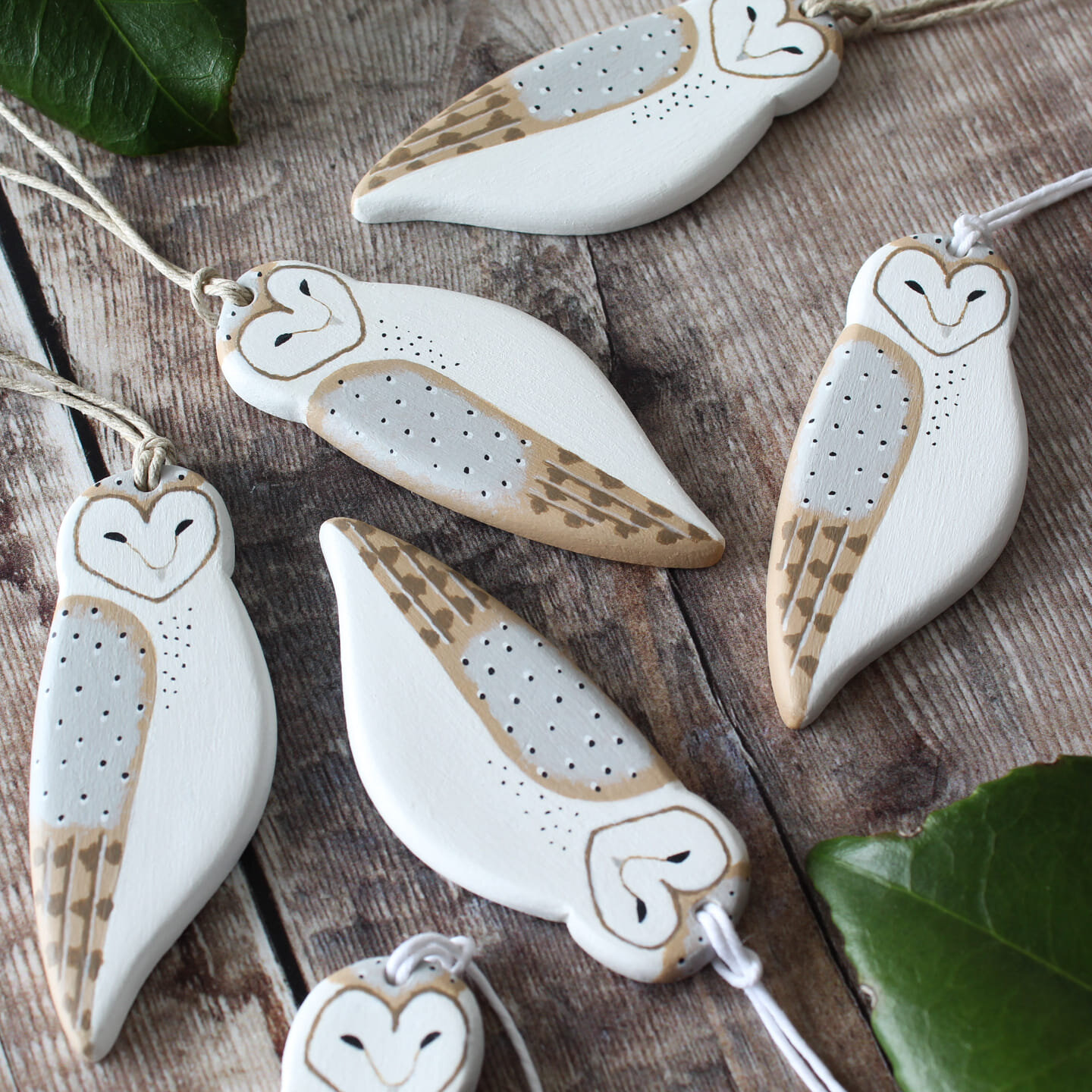 Five clay owl hanging ornaments sit on a rustic wood table. They are painted white, with grey and brown wing accents