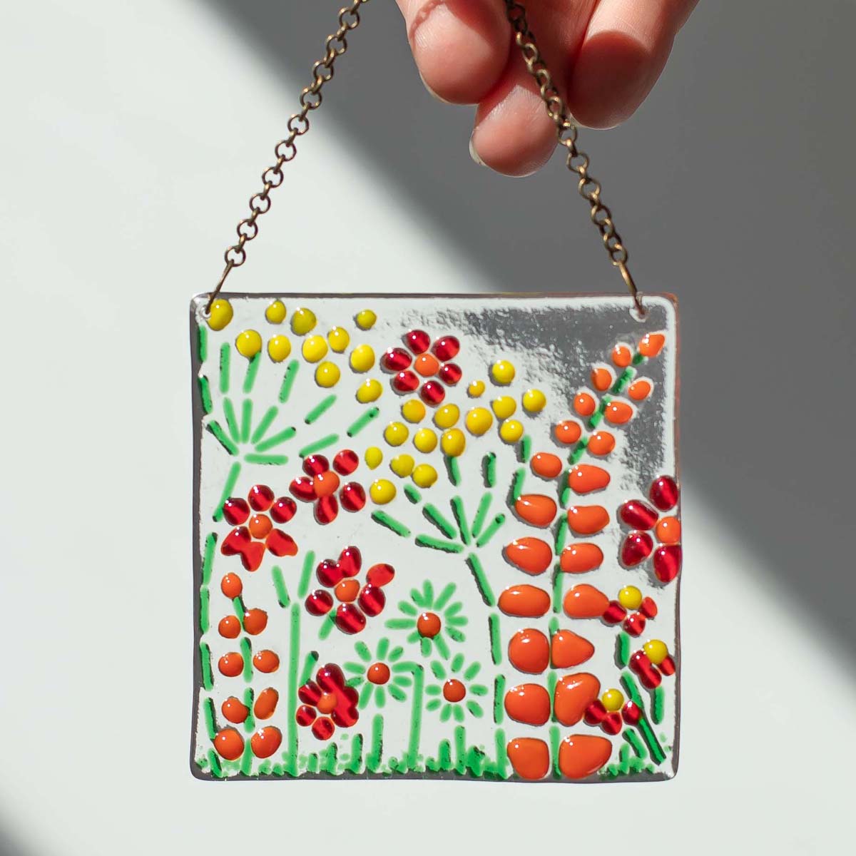 Hand holding a square fused glass hanger with red and orange flowers and green leaves