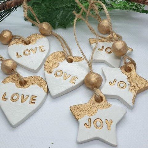 Clay ornaments sit on top of a white table. Three are love hearts and three are stars. They are painted white and gold in a Christmas theme, with Love and Joy painted on them.