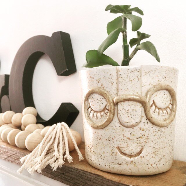 A white and brown speckled plant pot sits on top of a wooden mantle piece. The vase has painted on eyes that are closed, and glasses made from clay. It sits beside a wooden letter 'C' with some decorative wooden beads.