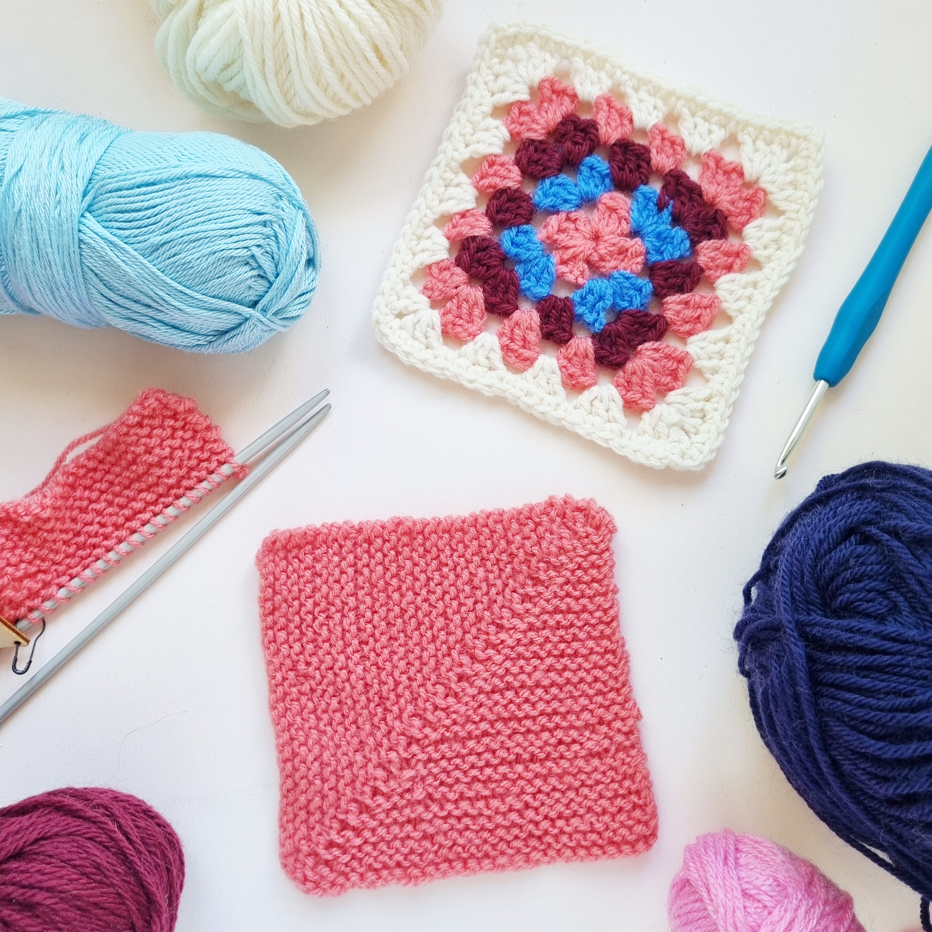 A pink knitted mitred square and a pink, blue and maroon crochet granny square sit on a table surrounded by yarn, knitting needles and a crochet hook