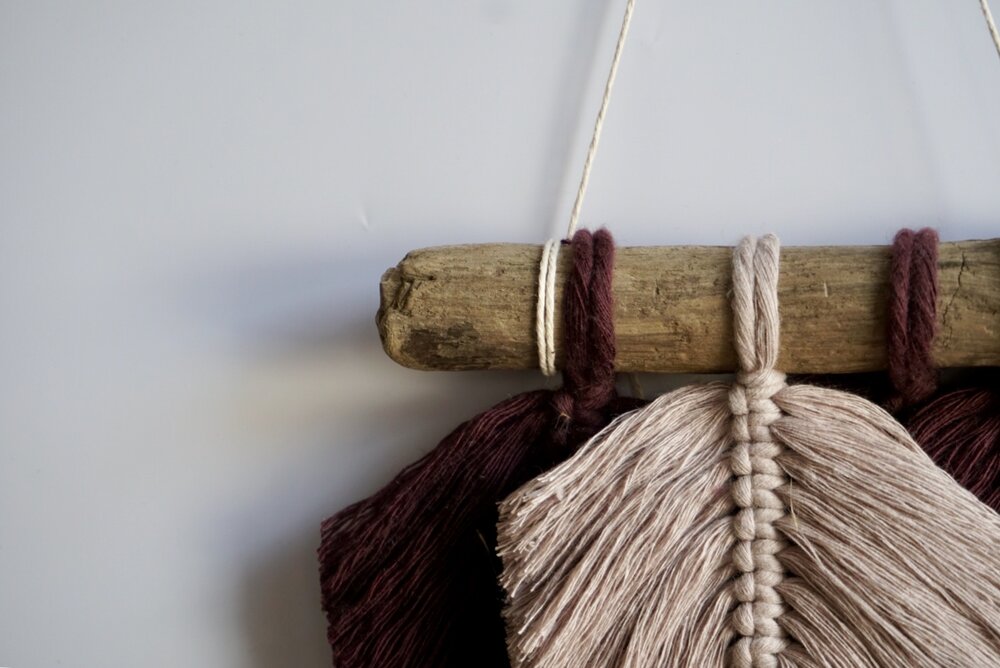 A close-up of three macrame feathers hanging on a piece of driftwood.