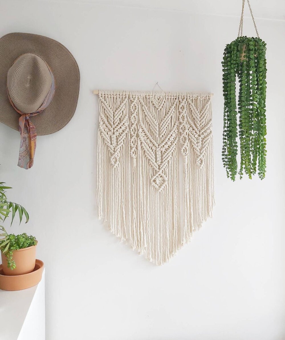 Image of a decorative macrame wall hanging. A summer hat hangs to the left, and a string of pearls hangs from the ceiling to the right.