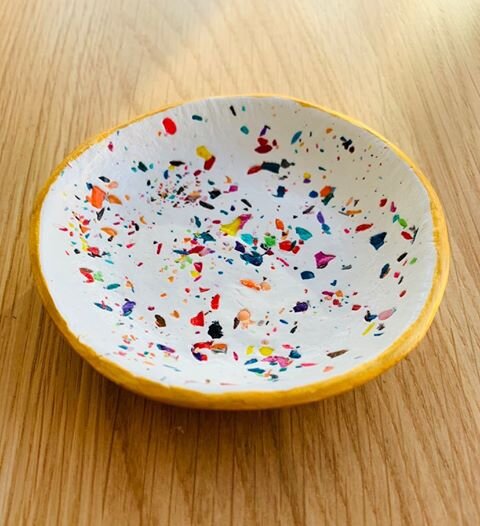 A small, round, shallow dish sits on top of a wooden table. The edges are painted yellow, with a multi-coloured terrazzo pattern in the middle.