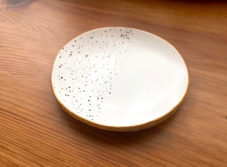 A small, round, shallow trinket dish sits atop a wooden table. It is painted white, with one half covered in tiny, black polka dots of different sizes.