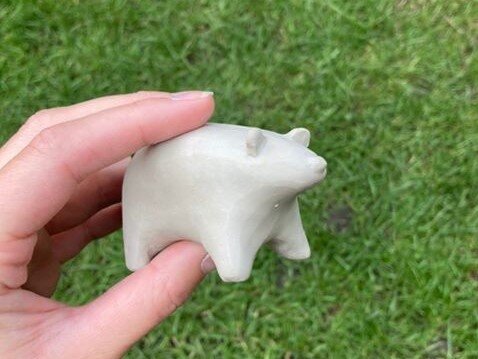 A hand-sized, white clay bear is held in frame above some green grass. It does not have any painted features.