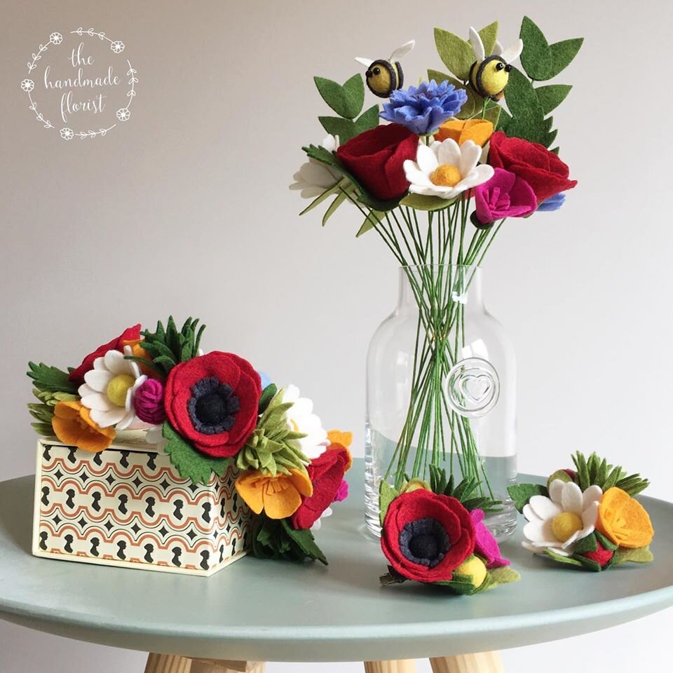 A bunch of felt flowers stand in a clear vase. Felt flowers lay decoratively across the table