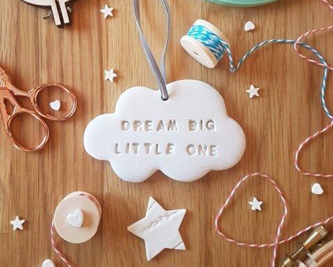 A white hanging ornament shaped like a cloud sits atop a wooden table surround by scissors, string, and tiny clay starts. It reads the words: "Dream big, little one".