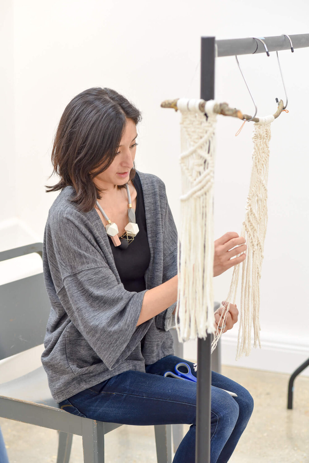 A woman sits working on a long macrame decoration.