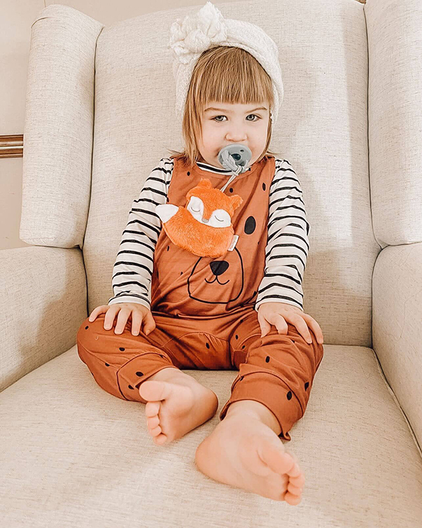 little girl wearing brown baby overalls and a fox soother