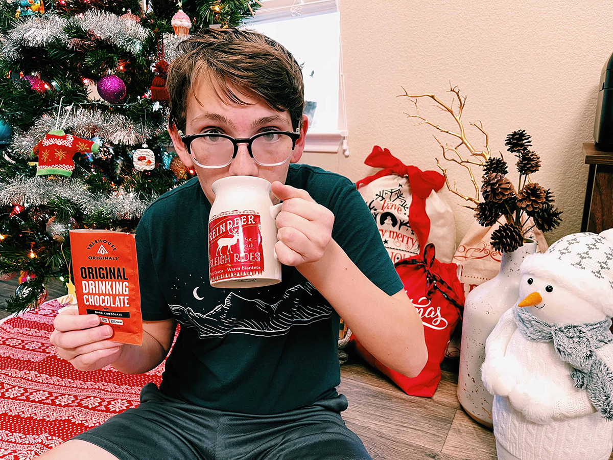 kid drinking hot cocoa next to christmas tree