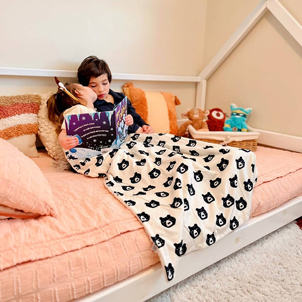 toddler girl reading a book with her brother under a plush bear print blanket