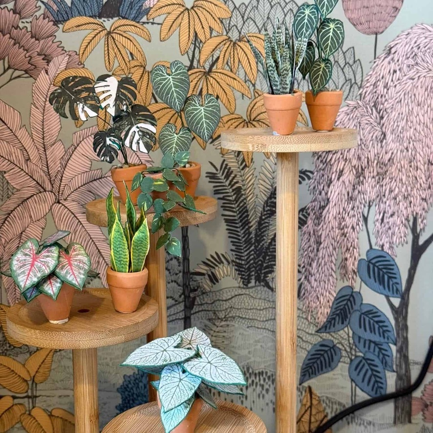 Mini paper plant display with illustrated jungle backdrop Miniature handmade paper plants in terracotta pots displayed on wooden pedestal stands in front of illustrated botanical wallpaper