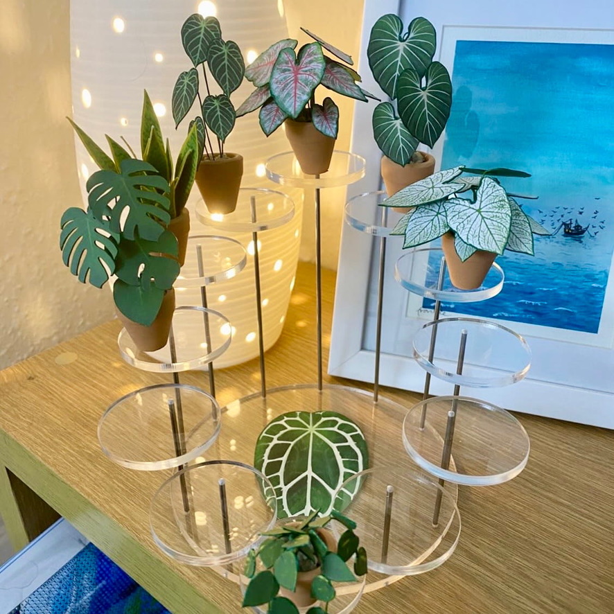 Mini paper plant collection on acrylic display stand Miniature handmade paper plants displayed on a clear tiered acrylic stand, lit by a white lamp with dotted lights