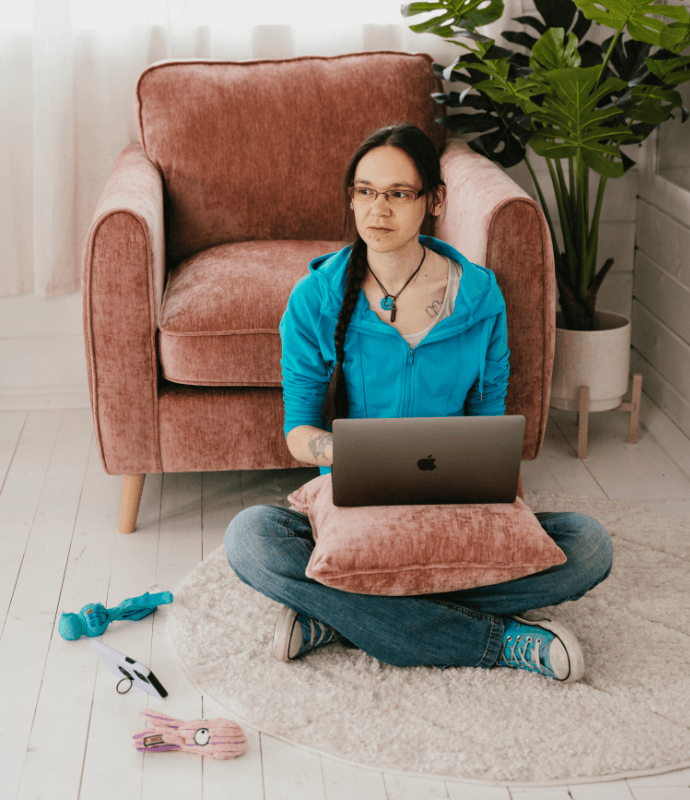 Photograph of caucasian woman sitting on floor with a laptop on a rose coloured cushion on her lap