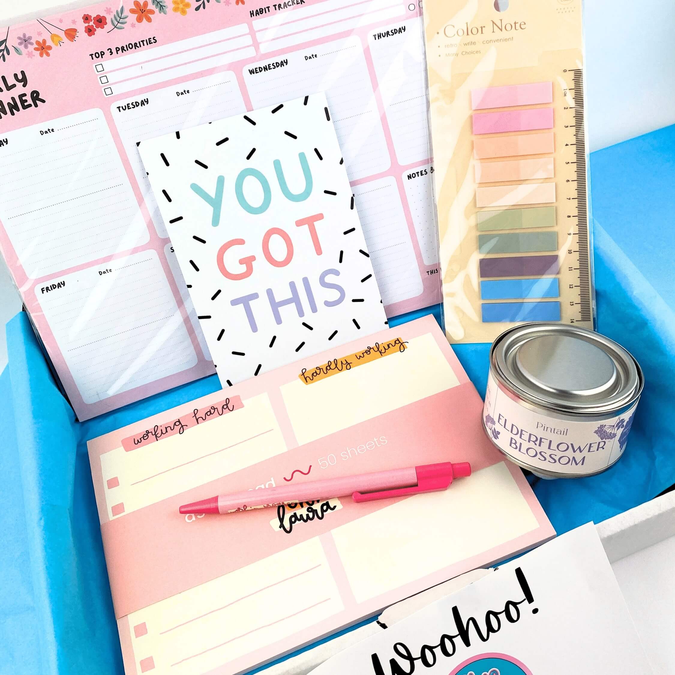 Colourful planner, notepad, sticky notes and candle in metal tin within a box with blue tissue paper