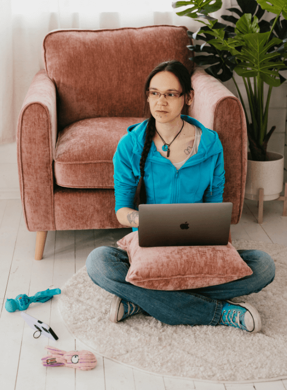 Jeni Machon Certified Social Media Manager Blue Raspberry Box Photograph of Jeni Machon sitting on floor with a laptop on a rose coloured cushion on her lap