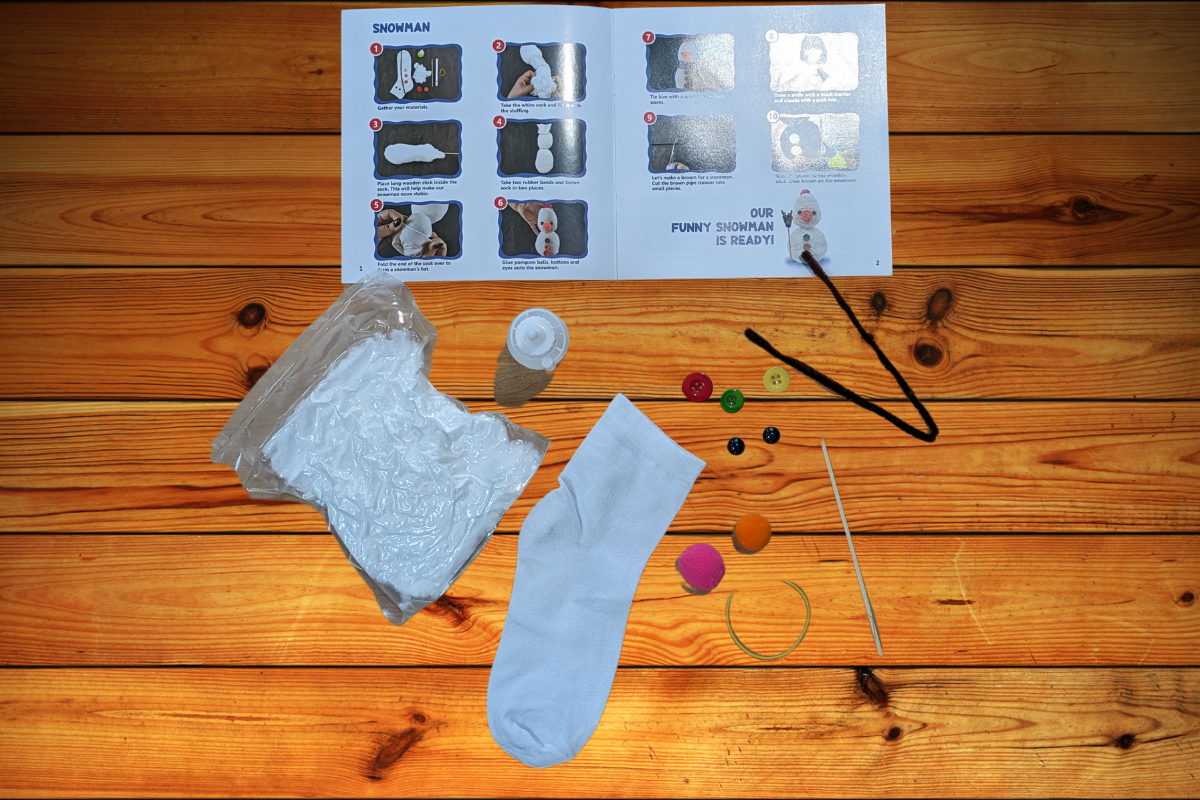 Materials for a snowman craft are laid out on a wooden surface. Items include a white sock, cotton stuffing, colorful buttons, a small bottle, embroidery thread, a rubber band, a pink felt circle, an orange felt triangle, googly eyes, and an instruction sheet detailing the steps—perfect for your Kr8tive Kids Craft Subscription Box from Unboxing the Bizarre.