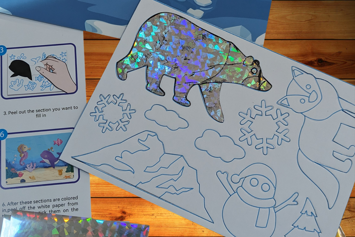 A Kr8tive Kids Craft Subscription Box by Unboxing the Bizarre featuring a holographic polar bear, snowflakes, and other winter-themed cutouts on a white sheet. Nearby are instructions with drawings on how to complete the project. The background includes a wooden surface and additional bright holographic sheets.